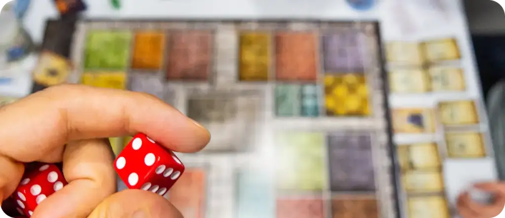 Közelkép a kártyákkal és színes rácsos táblával ellátott társasjáték felett piros kockát tartó kézről az asztalon