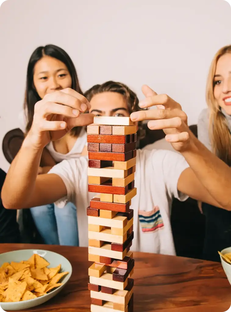 Barátok csoportja otthon fából készült építőelemekkel játszik, miközben az asztal körül harapnivalókat fogyasztva a toronyra koncentrálnak.