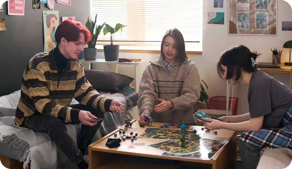 Három barát otthon, egy kényelmes nappaliban ülve, kártyákkal, miniatűrökkel és játéktáblával körülvéve, egy asztal körül ülve társasjátékot játszanak.